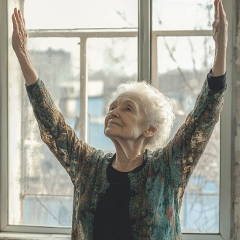 Жінка похилого віку в зручному одягу виконує позу Тадасана (гора) з витягнутими догори руками на тлі вікна